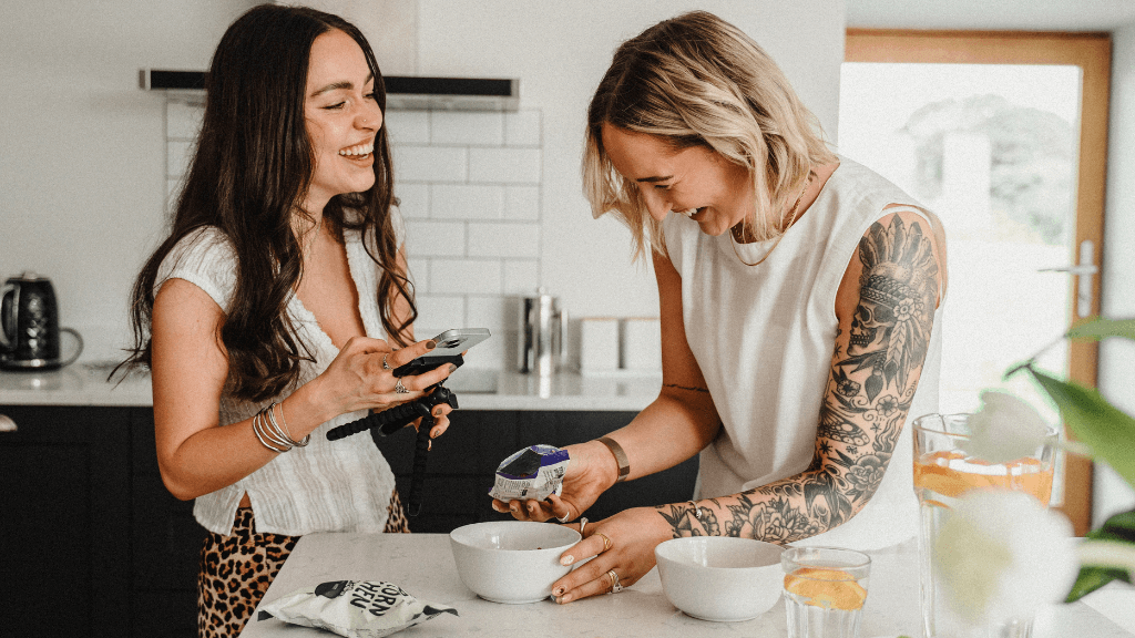 Two freelance social media managers creating content together in a bright kitchen, smiling while working on a food brand shoot