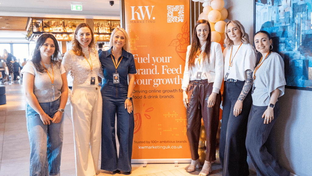 Team photo from FUEL LIVE 2026 showing the KW Marketing team standing together in front of an orange branded banner, smiling and wearing event lanyards at a busy venue.